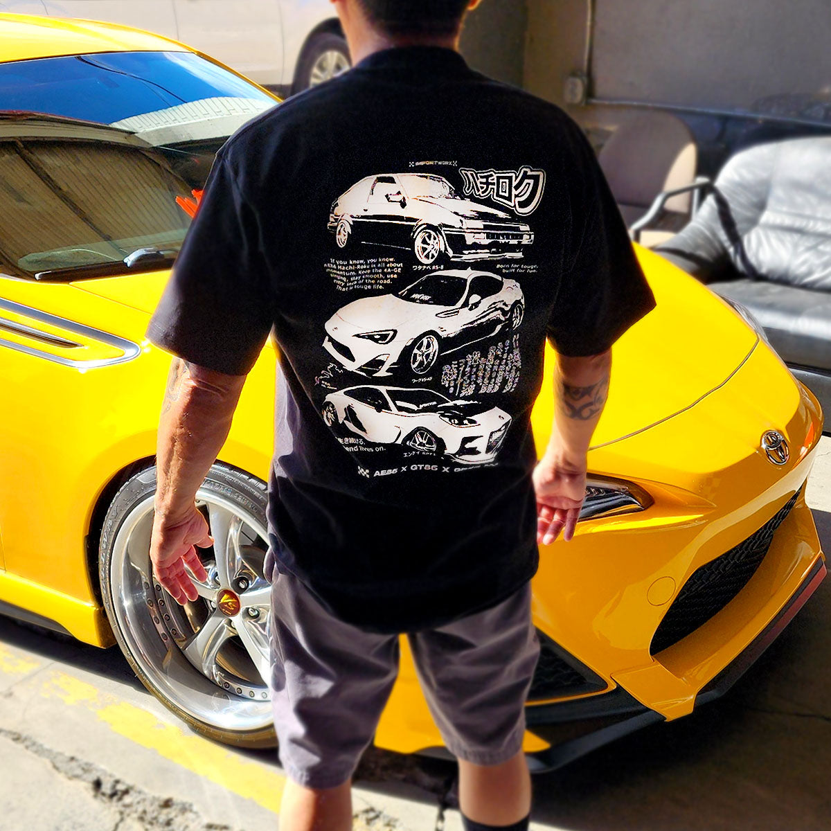 Person wearing a black ImportWorx t-shirt with AE86 ZN6 and GR86 car graphics standing next to a yellow sports car.